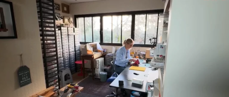 Artiste blonde en chemise claire assemblant une création à son plan de travail, entourée de casiers de rangement en atelier