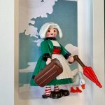 Figurine Bécassine en robe verte, coiffe blanche, valise et parapluie rouge, avec une oie, dans un cadre blanc