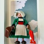 Figurine Bécassine en robe verte, coiffe et tablier blancs, valise brune et parapluie rouge, dans un cadre blanc
