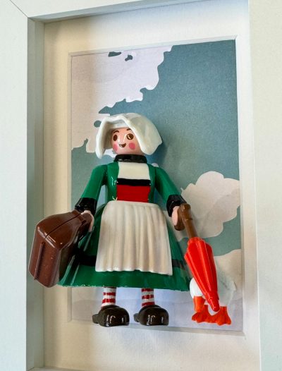 Figurine Bécassine en robe verte, coiffe et tablier blancs, valise brune et parapluie rouge, dans un cadre blanc