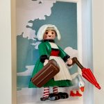 Figurine Bécassine en robe verte, coiffe blanche, valise, parapluie rouge et oie, dans un cadre blanc