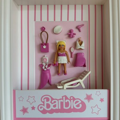 Tableau Boite poupée mannequin avec figurine Playmobil blonde, accessoires roses et logo Barbie dans un cadre blanc