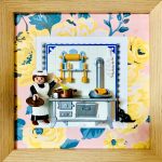 Tableau Désirée, figurine Playmobil de cuisinière en tablier blanc devant un fourneau ancien, chat noir et papier peint