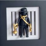 Tableau Garde à vous, figurine noire à casquette avec écharpe et clairon dorés sur fond rayé encadré de blanc