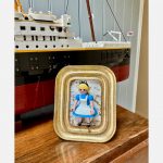 Tableau ALICE de Mlle Louise, figurine en robe bleue et tablier blanc dans un cadre doré, devant un paquebot en briques
