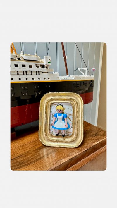 Tableau ALICE de Mlle Louise, figurine en robe bleue et tablier blanc dans un cadre doré, devant un paquebot en briques