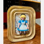 Tableau ALICE, figurine en robe bleue et tablier blanc dans un cadre doré posé sur une table en bois