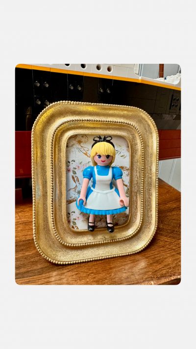 Tableau ALICE, figurine en robe bleue et tablier blanc dans un cadre doré posé sur une table en bois