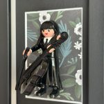 Figurine Playmobil Mercredi en robe noire tenant un violoncelle, encadrée sur fond floral blanc et vert