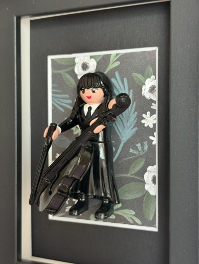 Figurine Playmobil Mercredi en robe noire tenant un violoncelle, encadrée sur fond floral blanc et vert
