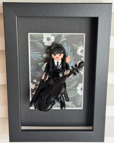 Tableau MERCREDI, figurine Playmobil en robe noire jouant du violoncelle sur fond floral, dans un cadre noir