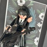 Figurine Mercredi en robe noire tenant un violoncelle noir devant un fond floral, detail du tableau MERCREDI