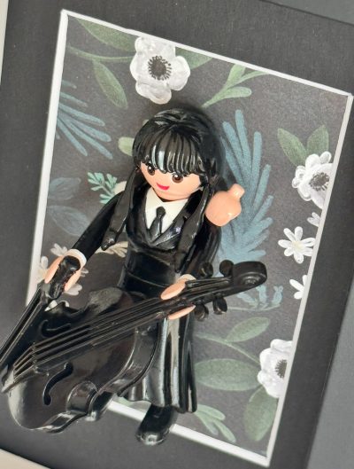 Figurine Mercredi en robe noire tenant un violoncelle noir devant un fond floral, detail du tableau MERCREDI