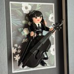 Tableau Mercredi, figurine Playmobil en robe noire jouant d'un violoncelle noir sur fond fleuri, cadre gris
