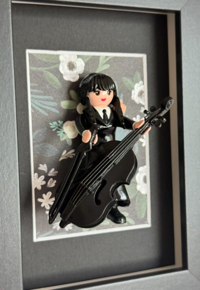 Tableau Mercredi, figurine Playmobil en robe noire jouant d'un violoncelle noir sur fond fleuri, cadre gris