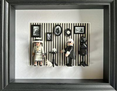 Tableau Rue Cambon avec deux figurines Playmobil, couturière en tailleur clair et créateur en noir, sur mur rayé