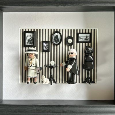 Tableau Rue Cambon avec deux figurines Playmobil, couturière en tailleur clair et créateur en noir, sur mur rayé