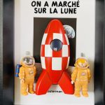 Deux figurines en combinaison spatiale orange avec casque transparent, posées de chaque côté d'une fusée rouge et blanche à damiers, dans un cadre noir avec un fond affichant le titre "On a marché sur la lune".
