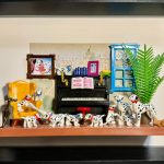 Scène miniature avec plusieurs dalmatiens de différentes tailles, un piano noir avec partitions, un fauteuil jaune, une fenêtre bleue et une plante verte, le tout encadré dans un cadre noir.