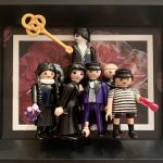 Figurines Playmobil représentant les membres de la famille Addams, habillés en noir avec des accessoires, dans un cadre noir avec un fond floral sombre.