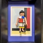 Figurine féminine avec cheveux noirs coiffés en chignon, portant une robe aux motifs géométriques et couleurs primaires (rouge, bleu, jaune, noir et blanc), encadrée sur un fond reprenant le style artistique de Piet Mondrian dans un cadre noir avec passe-partout bleu et blanc.