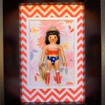 Figurine miniature inspirée de Wonder Woman, en costume rouge, bleu et or, tenant un lasso doré, encadrée dans un cadre noir avec un fond à motifs floraux et chevrons rouges et blancs.