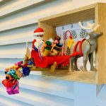 Figurine de Père Noël en costume rouge et blanc tenant une liste, assis dans un traîneau rouge rempli de cadeaux colorés, tiré par un renne gris avec un harnais rouge, encadré dans un cadre en bois avec un fond illustré de sapins de Noël.