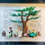 Scène avec une famille composée de deux adultes et deux enfants entourés d’animaux de la ferme (coq, poule, chien, biche, écureuil, sanglier) sous un grand arbre vert dans un cadre noir, fond illustré en noir et blanc.