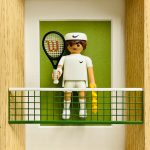 Figurine miniature de joueur de tennis représentant Roger Federer en tenue blanche avec bandeau, tenant une raquette noire avec logo rouge et une boîte de balles jaunes, derrière un filet vert, dans un cadre en bois avec fond vert.