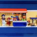 Scène miniature d’un stand de kebab intitulé "BLÜ KEBAB", avec plusieurs figurines : une femme assise à table buvant un verre, un homme en gilet jaune, une femme tenant un plateau, une petite fille, un vendeur derrière un comptoir bleu avec nourriture, un enfant jouant avec un chien, un couple âgé, et un homme barbu tenant une baguette, le tout sur un fond jaune encadré en bleu.