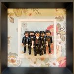 Cinq figurines en tenue noire brillante avec coiffures caractéristiques et symboles rouges sur les vêtements, dont une tient une guitare électrique noire, posées dans un cadre noir avec un fond floral coloré.