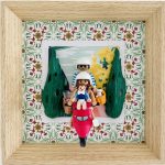 Trois figurines miniatures sur un scooter rouge ; un adulte en arrière-plan portant un casque blanc et lunettes noires, un enfant à vélo devant avec casque blanc, et une figurine assise tenant un panier avec un petit chien blanc, sur un fond décoratif floral vert et rouge avec un cadre en bois clair.