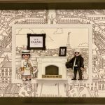 Deux figurines habillées en tenue élégante blanche et noire, posées dans un cadre noir avec un fond illustré représentant la ville de Paris et la Tour Eiffel, avec un cadre affichant "N°5 Chanel".