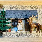 Figurine de fille en robe blanche et bleue avec un sac bleu, debout devant une petite maison en pierre avec un toit bleu, accompagnée d’un renne et d’un faon, à côté d’un sapin vert dans un cadre en bois avec un fond imprimé d’animaux de la forêt.