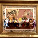 Douze figurines miniatures habillées en costumes d’époque disposées autour d’une longue table en bois avec divers objets sur la table, incluant des chandeliers dorés, dans un cadre en bois doré avec un fond fleuri rose et beige.