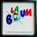 Deux figurines miniatures, un homme et une femme en blouse blanche, s’embrassant au centre d’un fond blanc portant le titre "LA BOUM" avec des lettres colorées (rouge, vert, bleu) rayonnant autour d’eux, accompagnées de plusieurs prénoms inscrits en cercle autour et en bas de l’image, le tout encadré dans un cadre noir.