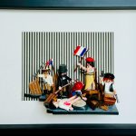Scène miniature inspirée de "La Liberté guidant le peuple" de Delacroix, avec plusieurs figurines dont une femme tenant un drapeau tricolore français, un homme en haut-de-forme, un soldat avec un fusil, plusieurs personnages en bonnet rouge avec armes, et une figure allongée au sol, le tout sur un fond à rayures verticales noires et blanches et encadré en noir.