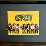 Huit figurines miniatures alignées devant un fond jaune avec le titre "BROOKLYN NINE-NINE", encadrées dans un cadre noir.