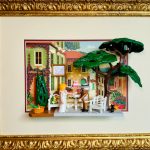 Deux figurines miniatures posées à une table blanche dans un jardin avec un grand arbre vert, décor floral et bâtiments jaunes avec volets verts en arrière-plan, le tout encadré dans un cadre doré orné et un fond beige.