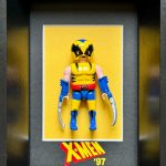 Figurine miniature de super-héros en costume jaune et bleu avec masque et griffes argentées, encadrée sur un fond jaune avec le texte "X-MEN ’97" en bas.
