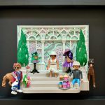 Trois figurines sur des marches devant une véranda décorée avec des guirlandes florales, deux top models de robes colorées sur mannequins, un cheval marron visible sur les côtés.