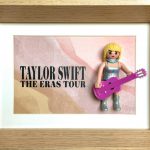 Figurine miniature blonde avec robe argentée et bottes argentées tenant une guitare rose vif, placée dans un cadre en bois à côté d’un fond imprimé avec le texte "Taylor Swift The Eras Tour".
