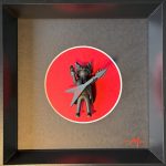 Figurine miniature noire avec des cornes de démon, tenant une guitare électrique en forme de V avec un logo rouge, posée au centre d’un cercle rouge dans un cadre noir avec une signature rouge "-M-" en bas à droite.