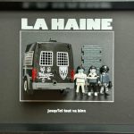 Trois figurines miniatures devant un mur en briques grises et à côté d’un fourgon noir de police avec inscriptions CRS, dans un cadre noir avec le titre "LA HAINE" en haut et la phrase "jusqu'ici tout va bien" en bas.