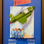 Quatre figurines miniatures habillées de combinaisons bleues avec des écharpes colorées (blanche, bleue, rouge, jaune) alignées devant un fond bleu clair avec un avion vert marqué "2" suspendu en haut, encadré dans un cadre noir avec le titre "Thunderbirds" en haut et des noms descriptifs en bas.