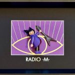 Figurine miniature en manteau violet, cheveux coiffés en pointes noires, tenant une guitare électrique noire en forme de V, posée devant un fond violet illustré d’un grand œil jaune stylisé avec des rayons blancs, encadrée dans un cadre noir.