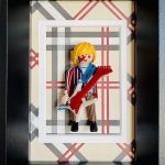 Figurine miniature avec cheveux blonds coiffés en boule, jouant de la guitare électrique blanche avec manche rouge, portant un costume rayé rouge, blanc et bleu sur fond beige à carreaux rouges, blancs et gris, encadré dans un cadre noir avec un passe-partout blanc.