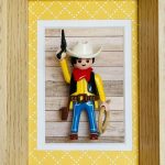Figurine miniature de cowboy avec chapeau blanc, foulard rouge, gilet noir, pantalon bleu, tenant un pistolet levé et un lasso, posée devant un fond imitation bois dans un cadre en bois clair avec un passe-partout jaune à motifs blancs.
