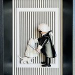 Figurine miniature en costume noir avec perruque blanche et masque noir, tenant une loupe et levant un drap blanc couvrant un petit chat blanc, le tout présenté dans un cadre noir avec un fond rayé noir et blanc et un cadre blanc intérieur.