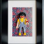 Figurine miniature avec cheveux noirs en coupe courte, lunettes rouges, nez de clown rouge, portant une salopette rayée bleue et blanche avec un t-shirt jaune, sur un fond décoré de cœurs rouges et motifs blancs sur fond bleu foncé.